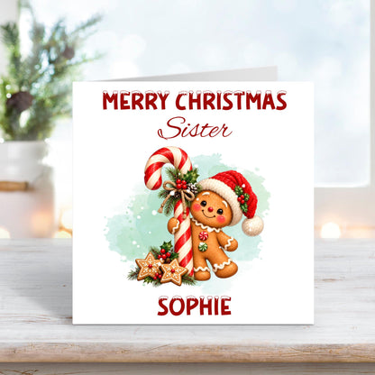 Personalised Merry Christmas Sister card featuring an adorable smiling gingerbread character wearing a red Santa hat, standing beside a large red and white candy cane decorated with holly, berries and pinecones. Festive gingerbread biscuits, soft mint green watercolour background and custom name Sophie.