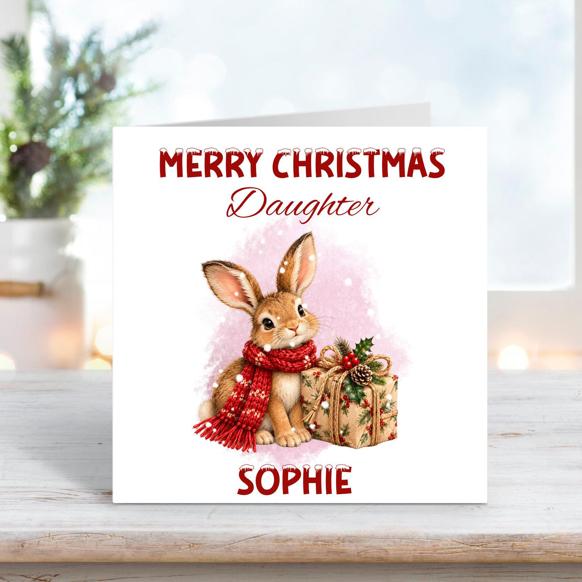 Personalised Merry Christmas Daughter card featuring an adorable brown woodland bunny wearing a cosy red knitted scarf, sitting beside a beautifully wrapped Christmas present decorated with holly, berries and a pinecone. Soft pink snowy watercolour background with custom name Sophie.