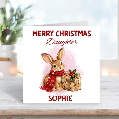 Personalised Merry Christmas Daughter card featuring an adorable brown woodland bunny wearing a cosy red knitted scarf, sitting beside a beautifully wrapped Christmas present decorated with holly, berries and a pinecone. Soft pink snowy watercolour background with custom name Sophie.