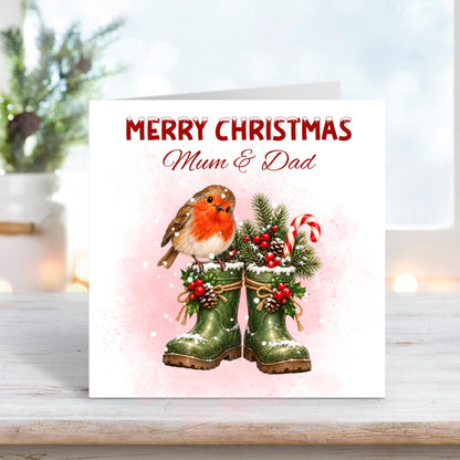 Merry Christmas Mum and Dad greeting card featuring a cute red-breasted robin perched on snowy green wellington boots filled with festive pine branches, holly berries, pinecones and a red and white candy cane. Soft pink watercolour background with falling snow.