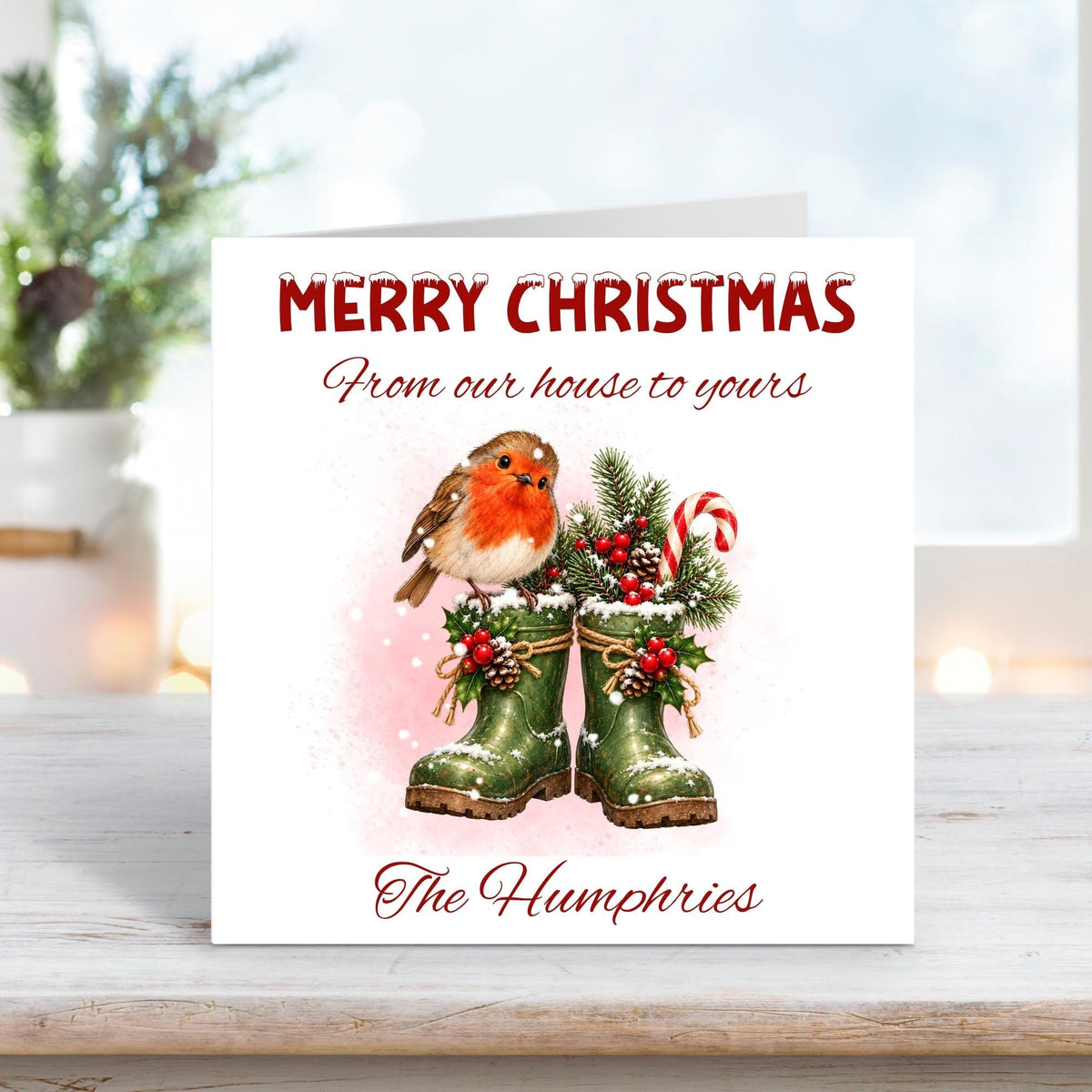 Personalised family Christmas card featuring a charming red-breasted robin perched on green wellington boots filled with festive pine branches, holly, berries, pinecones and a candy cane. Soft pink snowy watercolour background with the greeting “From our house to yours” and personalised family name “The Humphries”.
