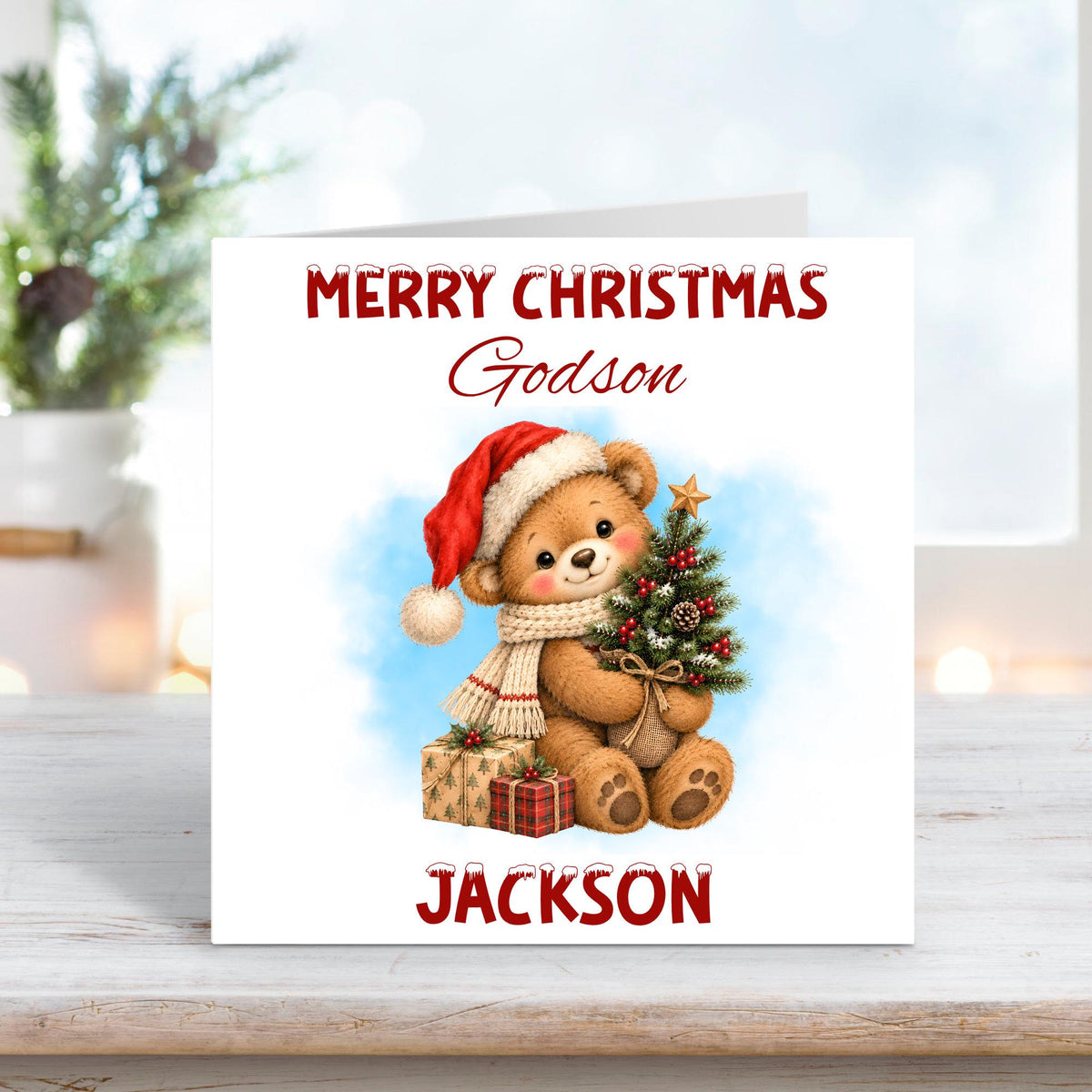 Personalised Merry Christmas Godson card featuring an adorable brown teddy bear wearing a red Santa hat and cosy cream knitted scarf, holding a miniature decorated Christmas tree with a gold star, red berries and pinecones. Wrapped presents, soft blue winter background and custom name Jackson.